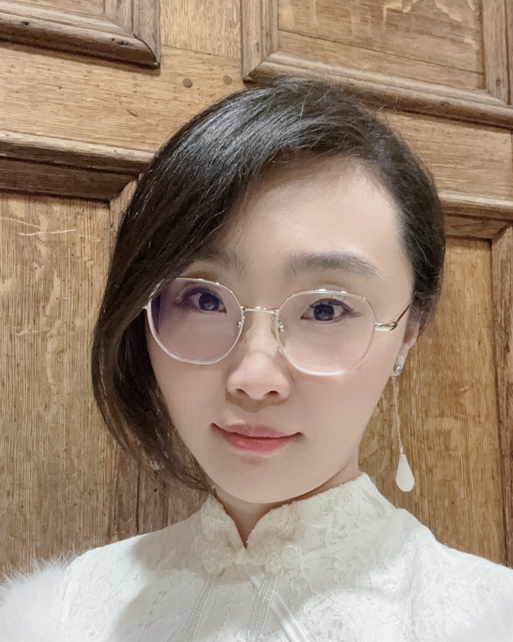 A woman with straight dark hair, wearing round glasses and a white lace top, stands in front of a wooden door. She has a single white drop earring and a calm expression on her face.