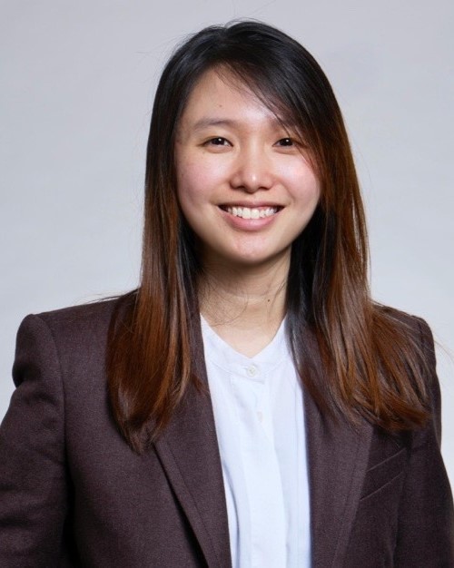 A woman with straight, long brown hair wears a dark blazer over a white shirt and smiles at the camera against a plain light background.