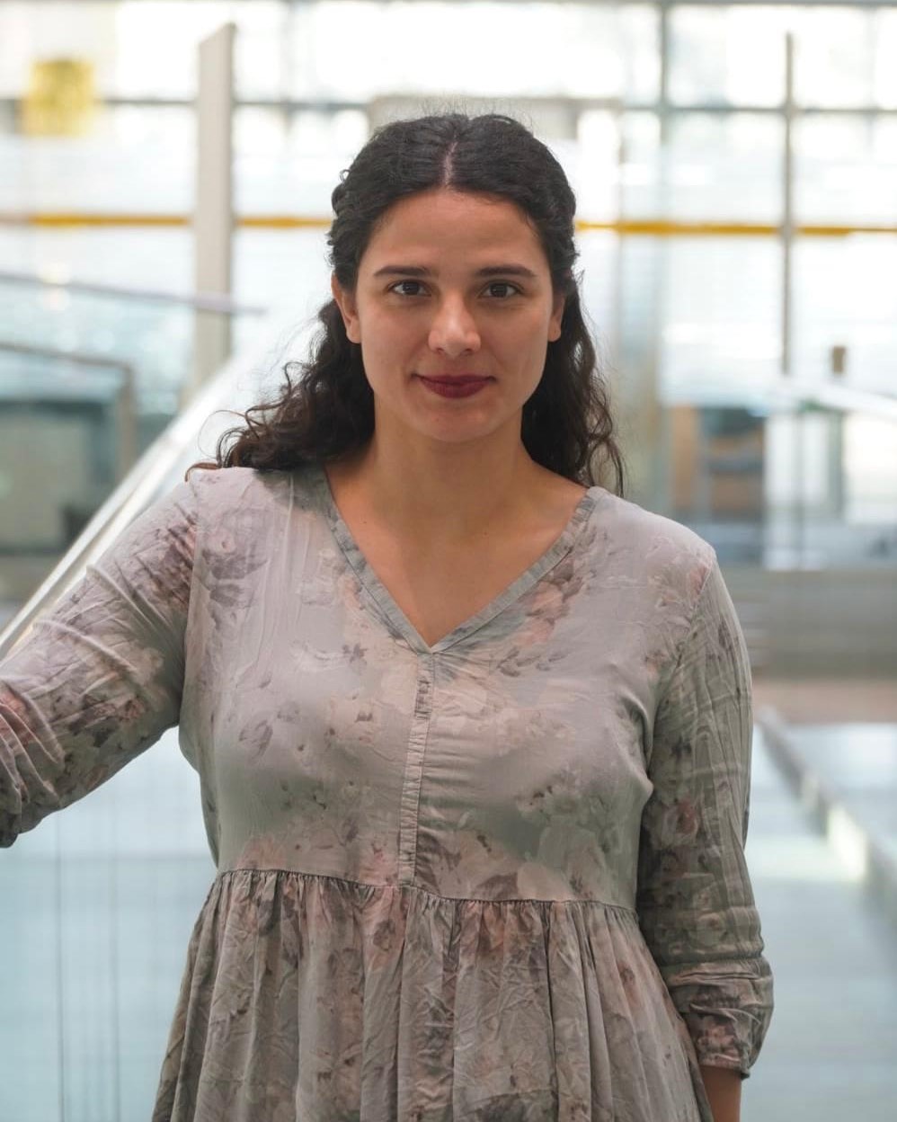 A woman with long dark hair, wearing a light gray floral dress, stands indoors near a glass railing, looking at the camera with a neutral expression. The background is bright and blurred.