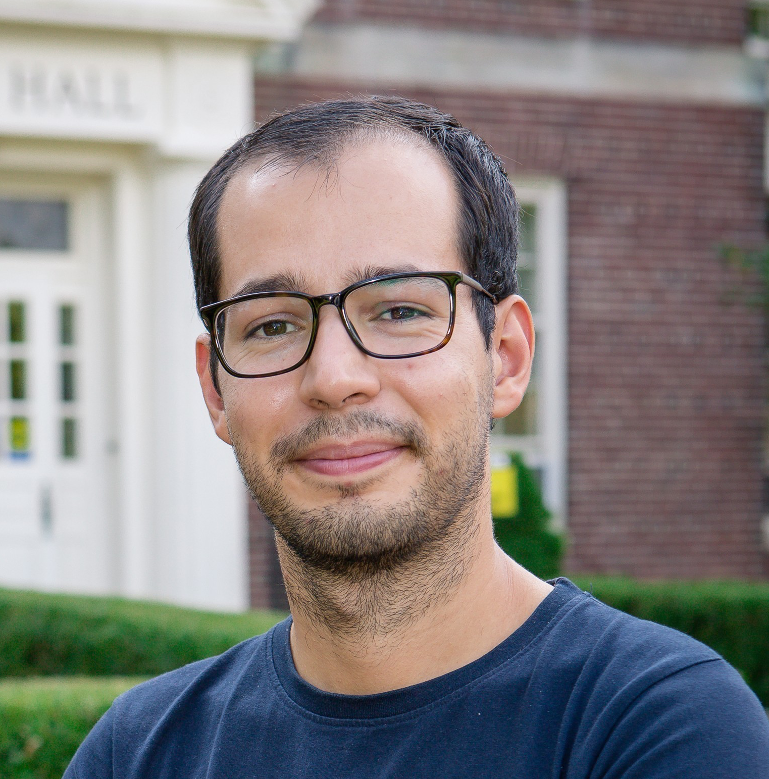 A man with short dark hair, glasses, and a trimmed beard is smiling slightly. He is wearing a navy blue t-shirt and standing outside in front of a brick building with white trim and greenery.