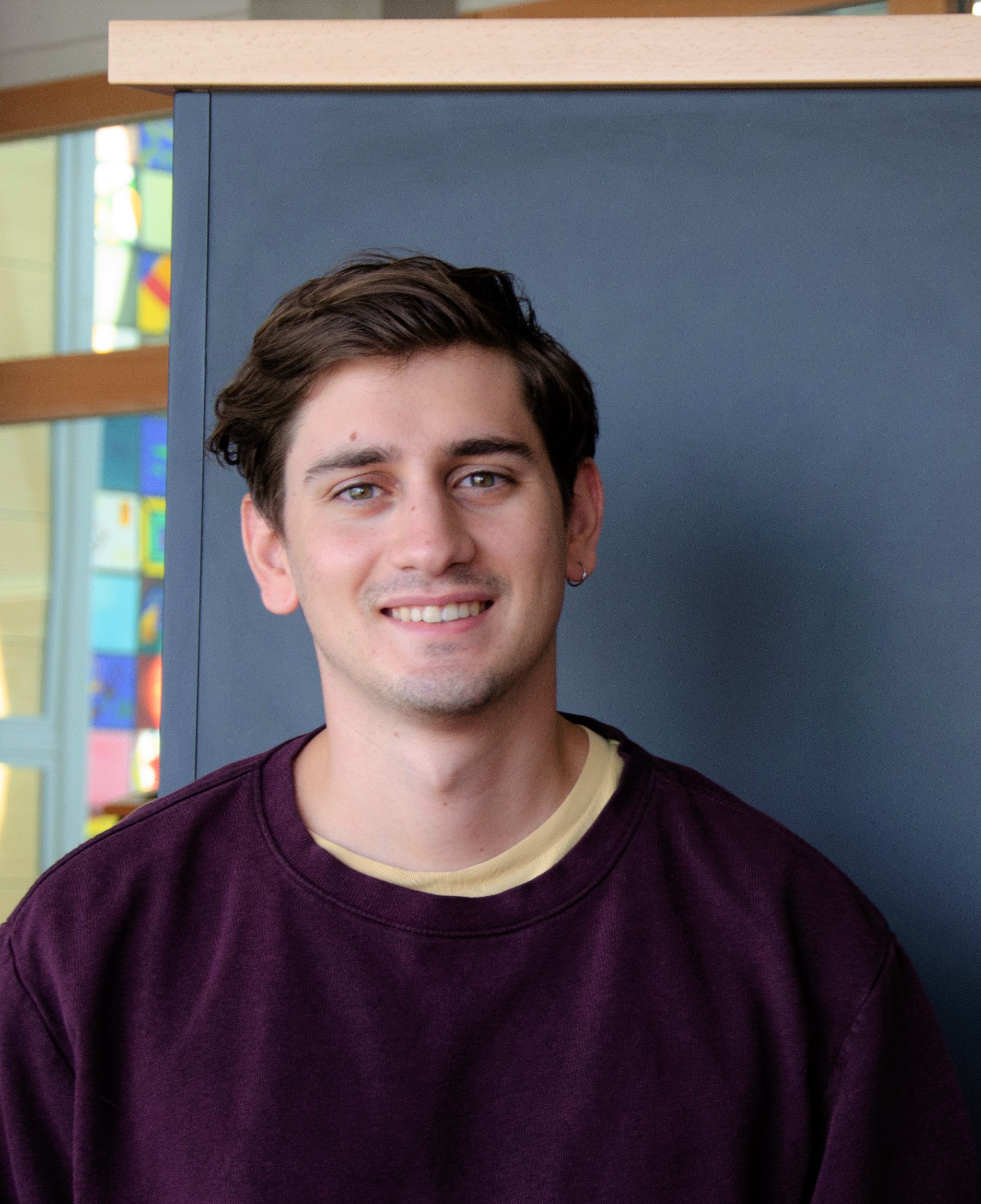 A young man with wavy brown hair and light skin, wearing a maroon sweatshirt over a beige t-shirt, smiles at the camera. He is standing indoors in front of a dark blue panel with colorful windows in the background.