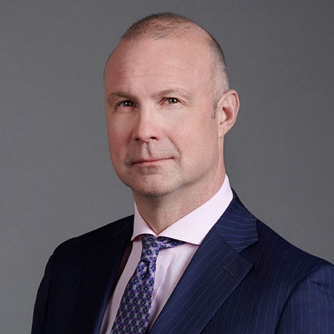 A middle-aged man with short, closely-cropped hair wearing a dark pinstripe suit, light pink shirt, and patterned tie, posing against a plain gray background.