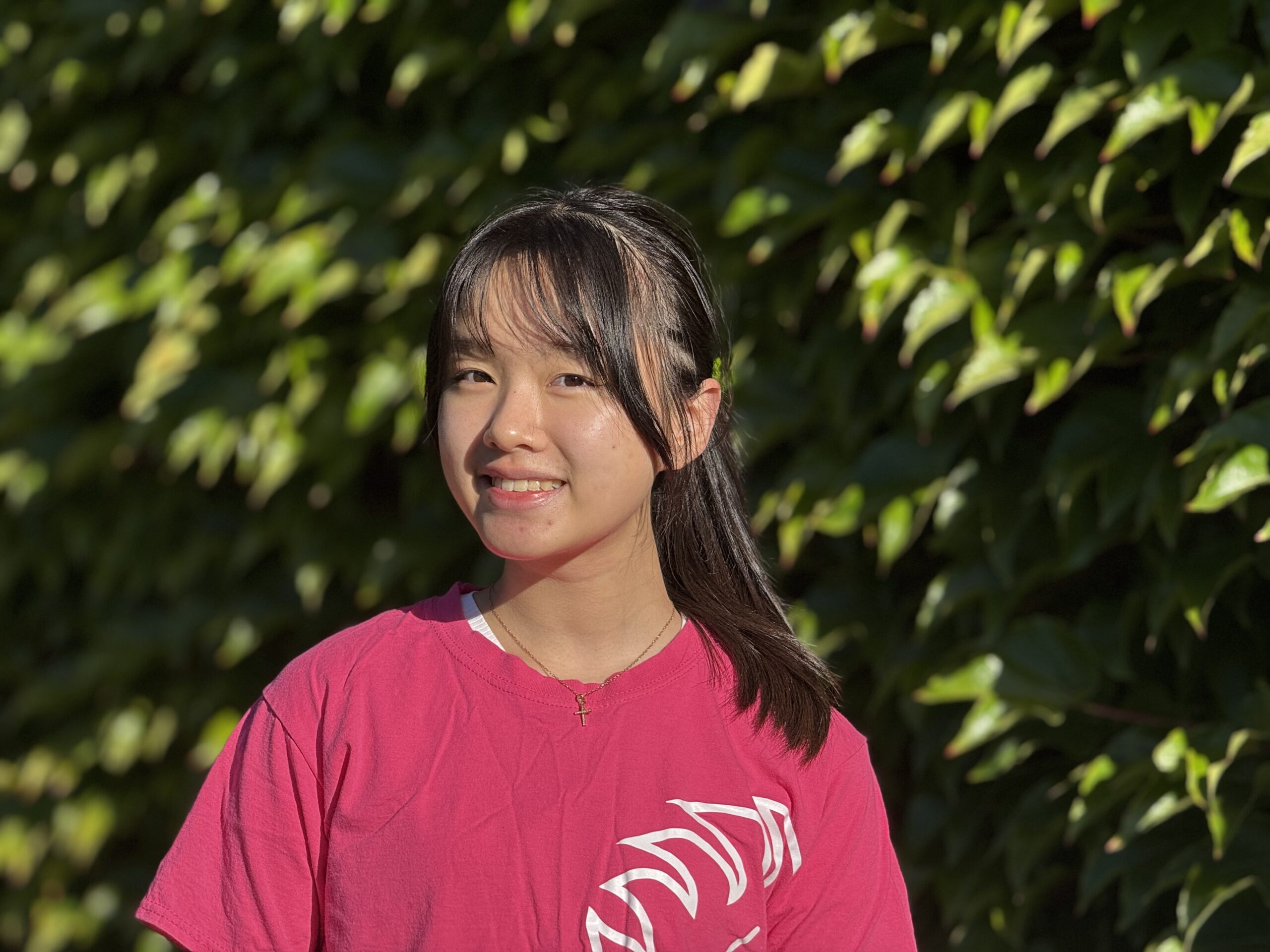 A female student with a pony tail wearing a pink Churchill t-shirt is standing in front of an ivy wall, smiling at the camera