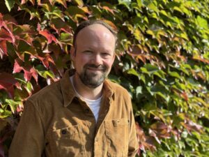 A man with a beard smiles at the camera. He is wearing a light brown coloured open neck shirt and he is standing in front of the Boston Ivy wall outside Churchill College