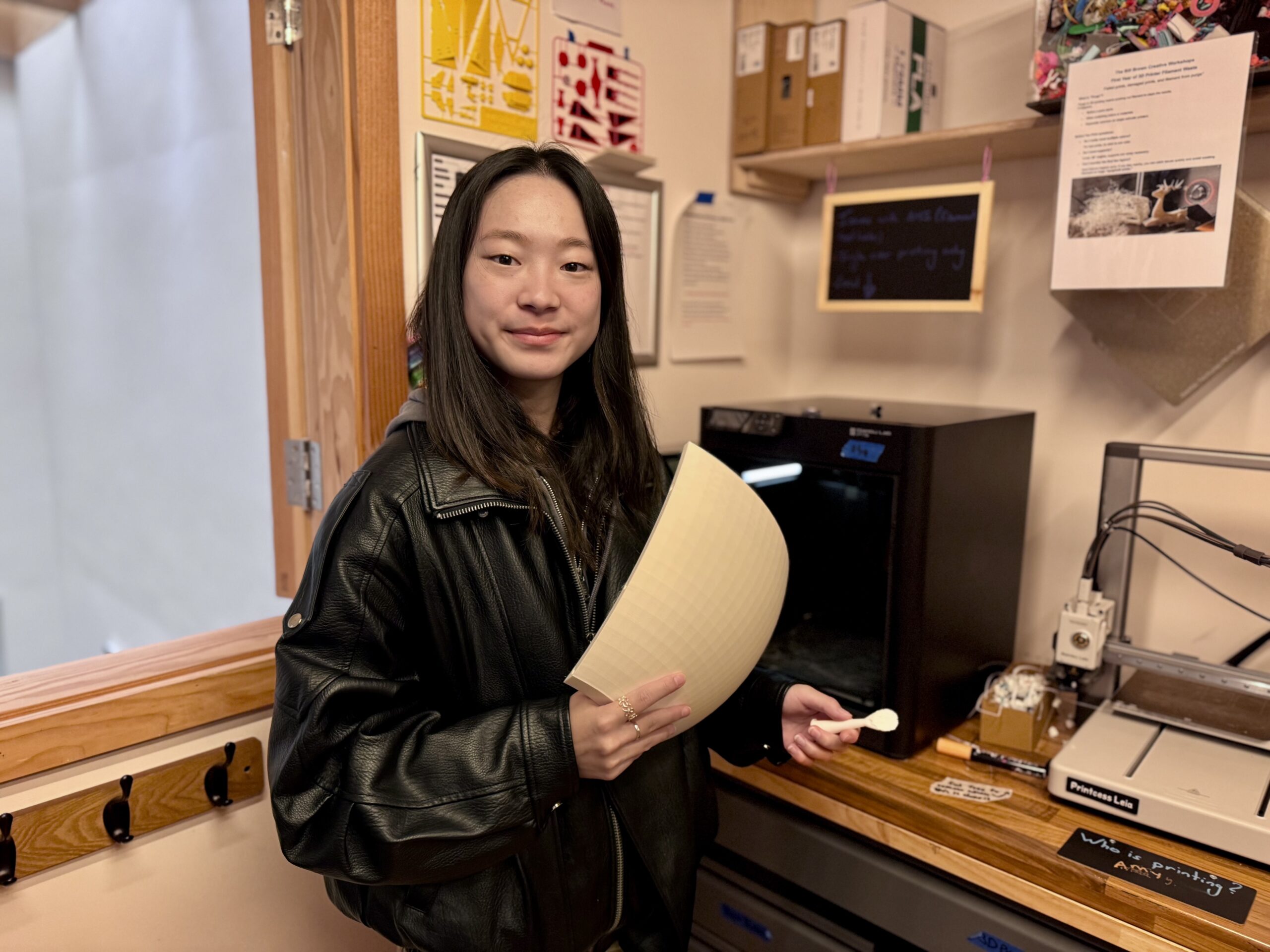 A young woman in a black leather jacket stands in a workshop, holding a sheet of material and a tool, with a 3D printer and various equipment and notes visible in the background.