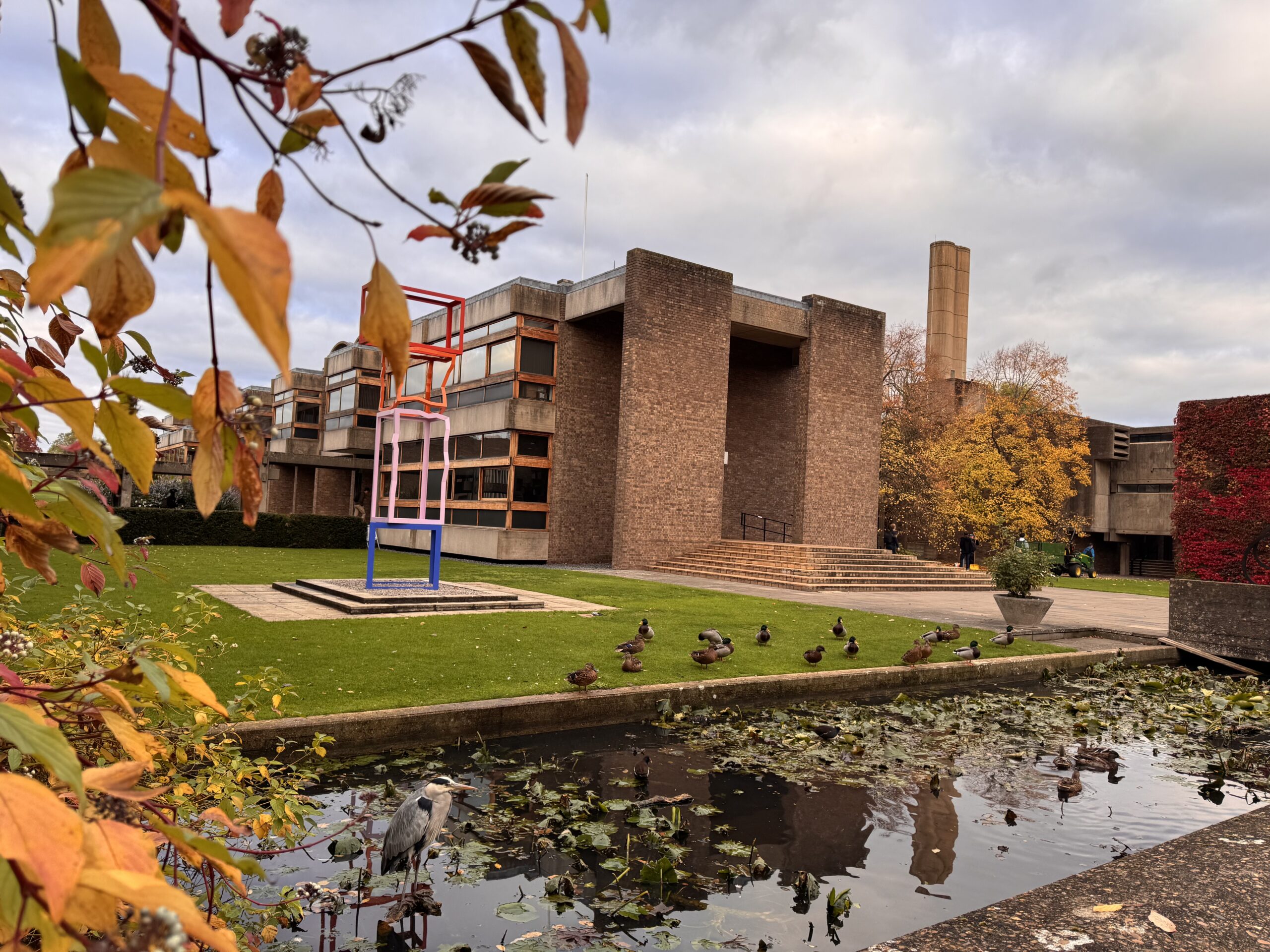 A modern brick building with large windows, surrounded by autumn trees and a grassy area with a colorful abstract sculpture. In the foreground, ducks and a heron are by a pond with lily pads.