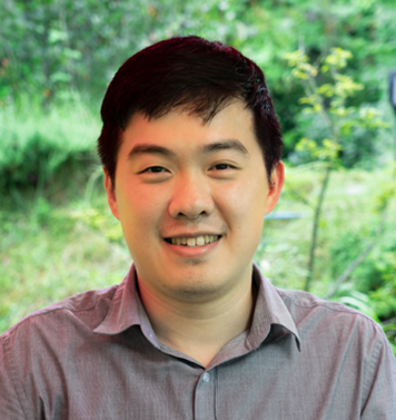 A young man with short dark hair, wearing a light gray collared shirt, smiles while sitting outdoors with lush greenery blurred in the background.
