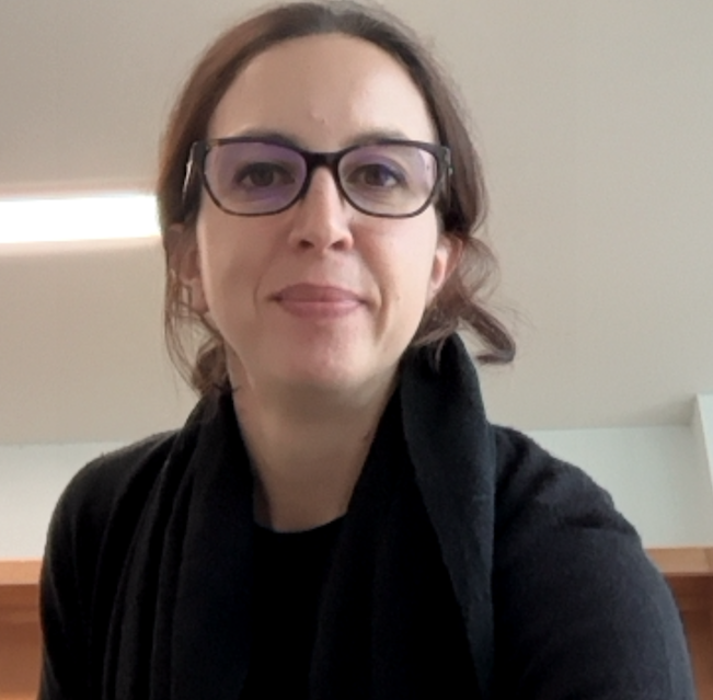 A woman with brown hair pulled back, wearing glasses and a black top, smiles softly at the camera indoors with a plain background.