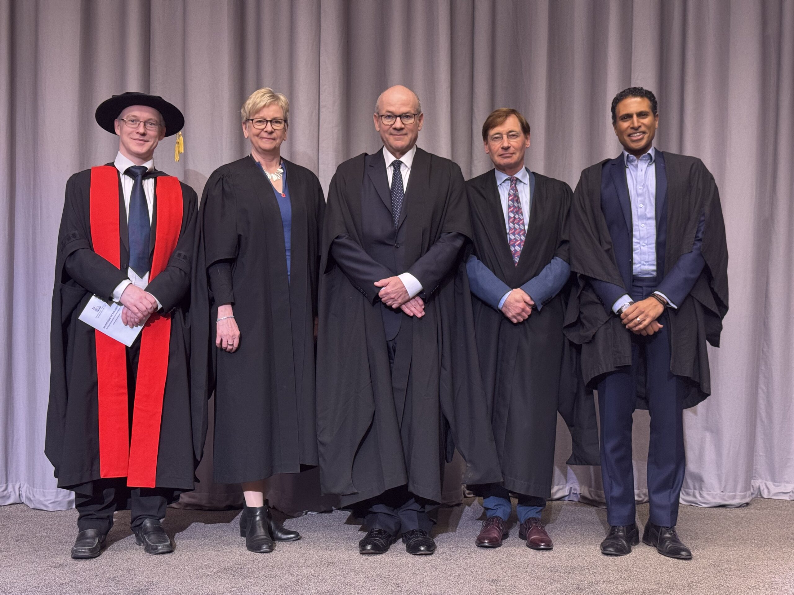 Five people in academic gowns and formal attire stand side by side in front of a gray curtain. One person wears a red and black robe with a hat, while the others wear black robes over business clothing.