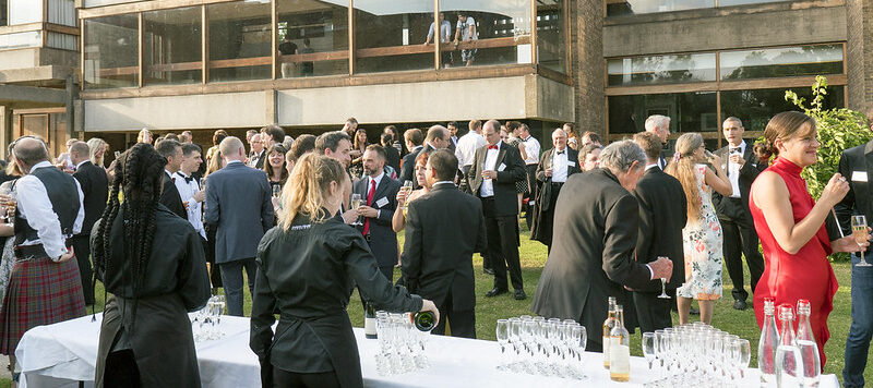 A large group of people dressed in formal attire socialize outdoors on grass near a modern building, with several people holding drinks and servers attending a white-clothed bar table with glasses and bottles.