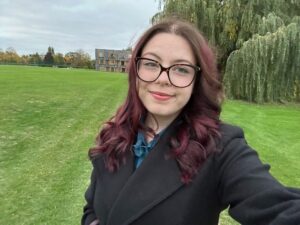 A girl with shoulder length hair and glasses is standing on the grass outside Churchill College. She is smiling at the camera.