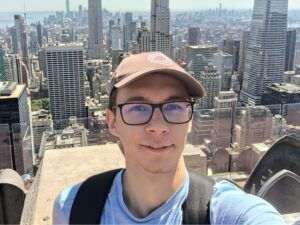 A selfie of Ivan wearing a baseball hat, glasses and casual t-shirt with the skyline of New York in the background