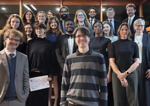 A group of 20 adults, dressed in formal and semi-formal attire, stand together indoors and smile at the camera; one person holds a certificate. The setting has wooden shelves and warm lighting.