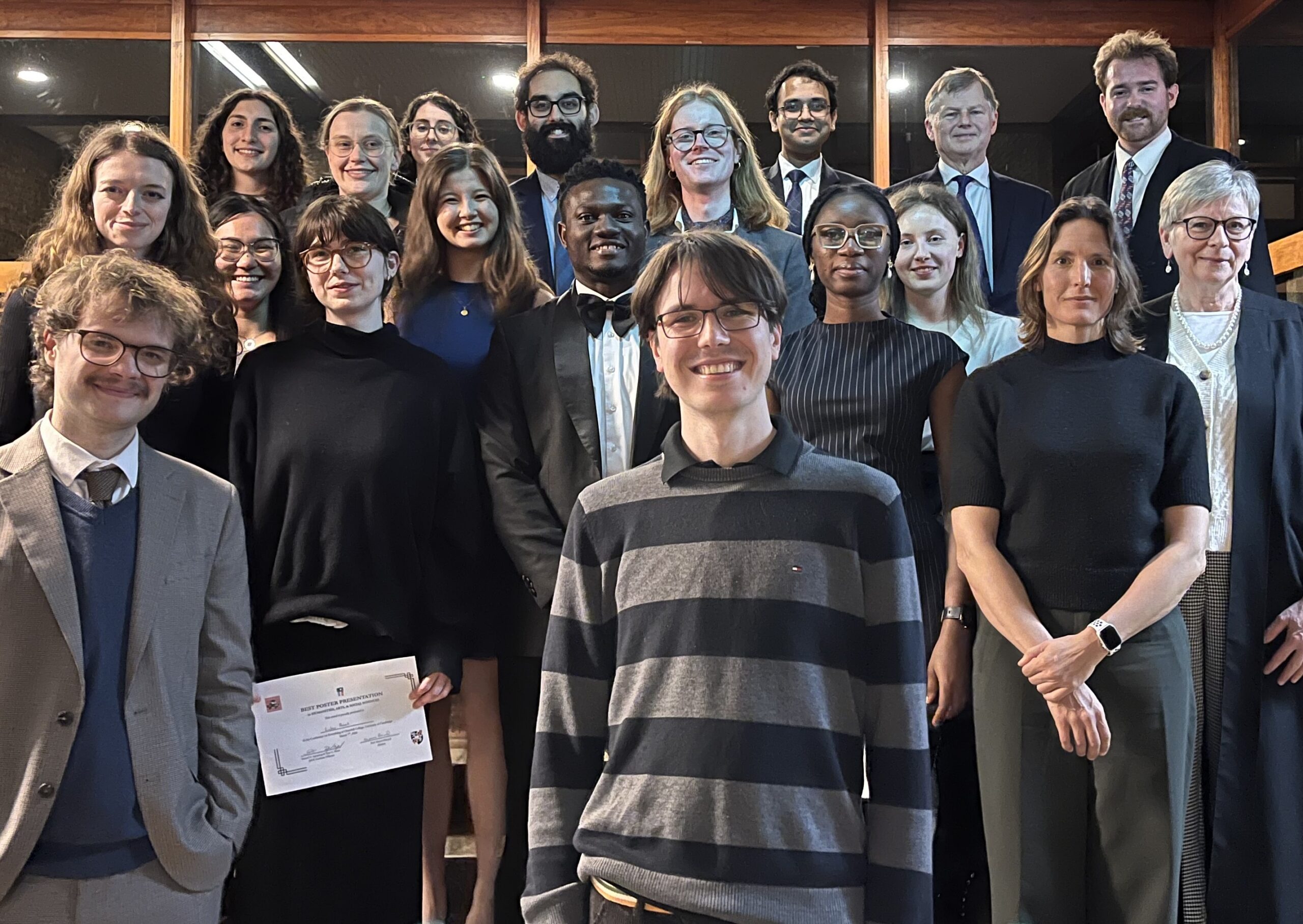 A group of 20 adults, dressed in formal and semi-formal attire, stand together indoors and smile at the camera; one person holds a certificate. The setting has wooden shelves and warm lighting.